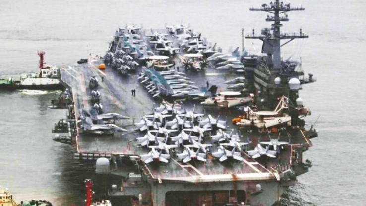 A Gerald R. Ford-class aircraft carrier under simulated attack by hypersonic missiles and drone swarms, symbolizing the "Carrier Killer" dilemma.